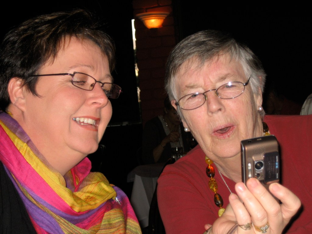 An older woman with grey hair and glasses is looking surprised as a younger person with dark hair and glasses shows them a mobile phone. 