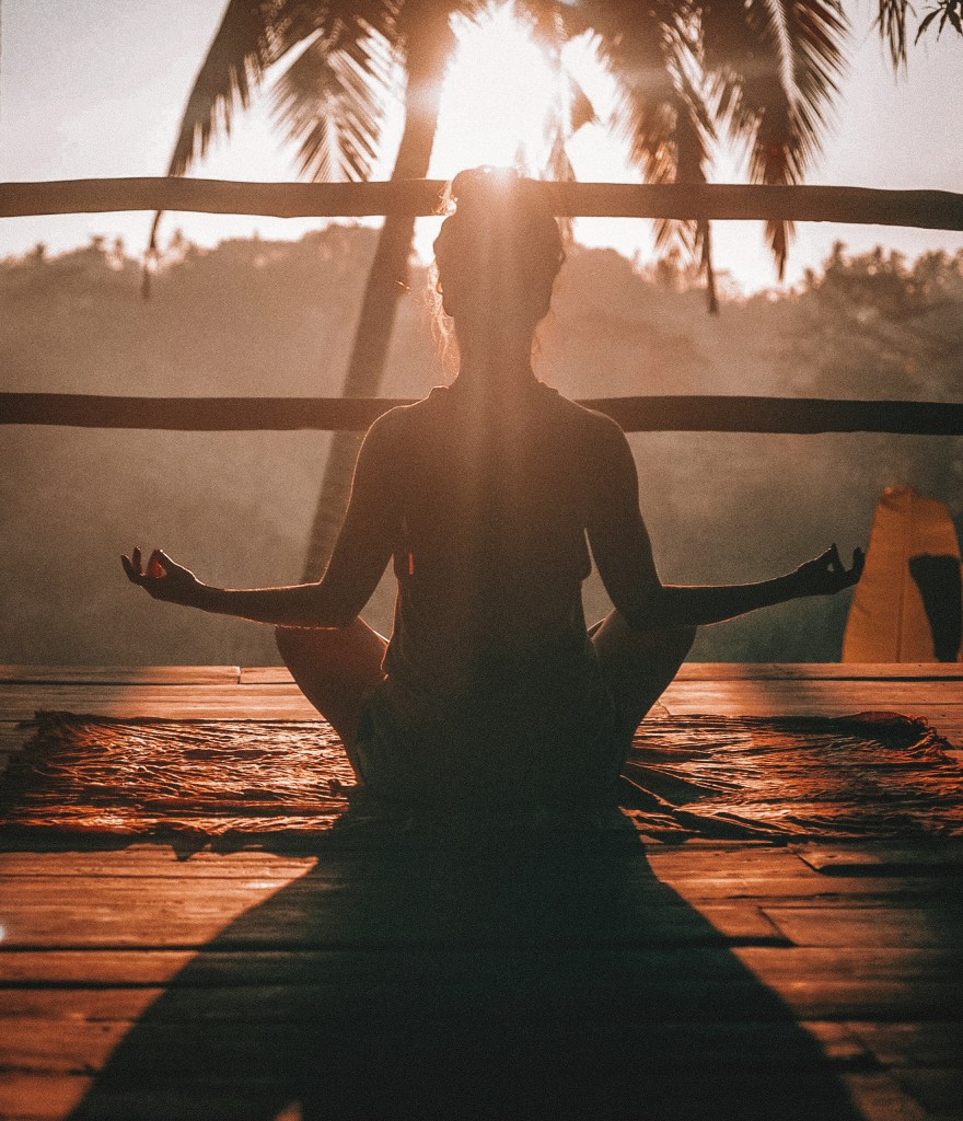 A young woman sits on a wooden deck in a meditation pose looking toward a palm tree with the sun behind it.